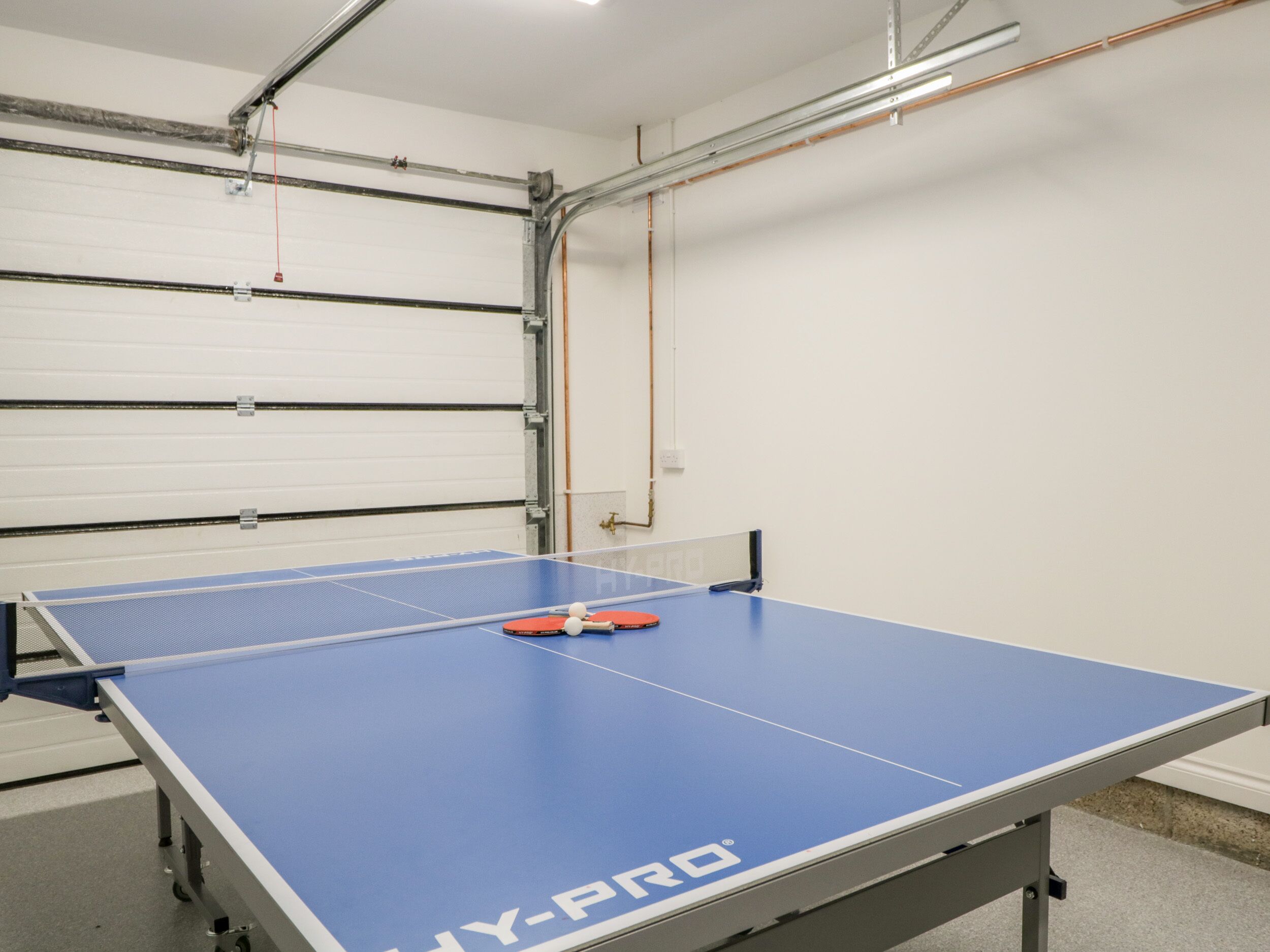 Ein Tischtennis-Tisch mit blauer Spielfläche und Netz in einem Raum mit weißen Wänden und einer Metall-Deckenkonstruktion.
