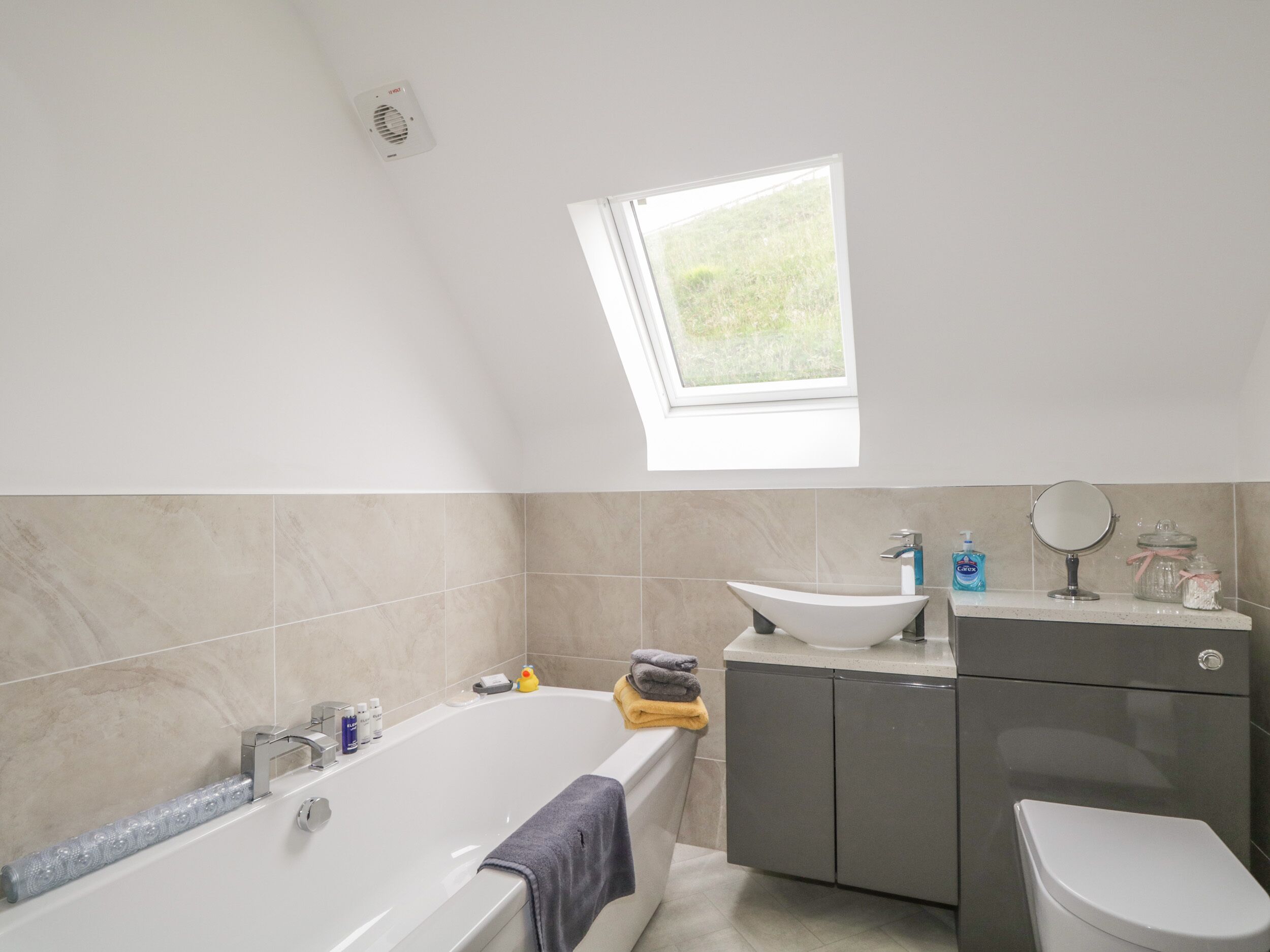 Ein modernes Badezimmer mit einer freistehenden Badewanne, einem Waschbecken und einer Toilette. Über dem Waschbecken befindet sich ein Oberlicht und über der Badewanne ein Fenster.