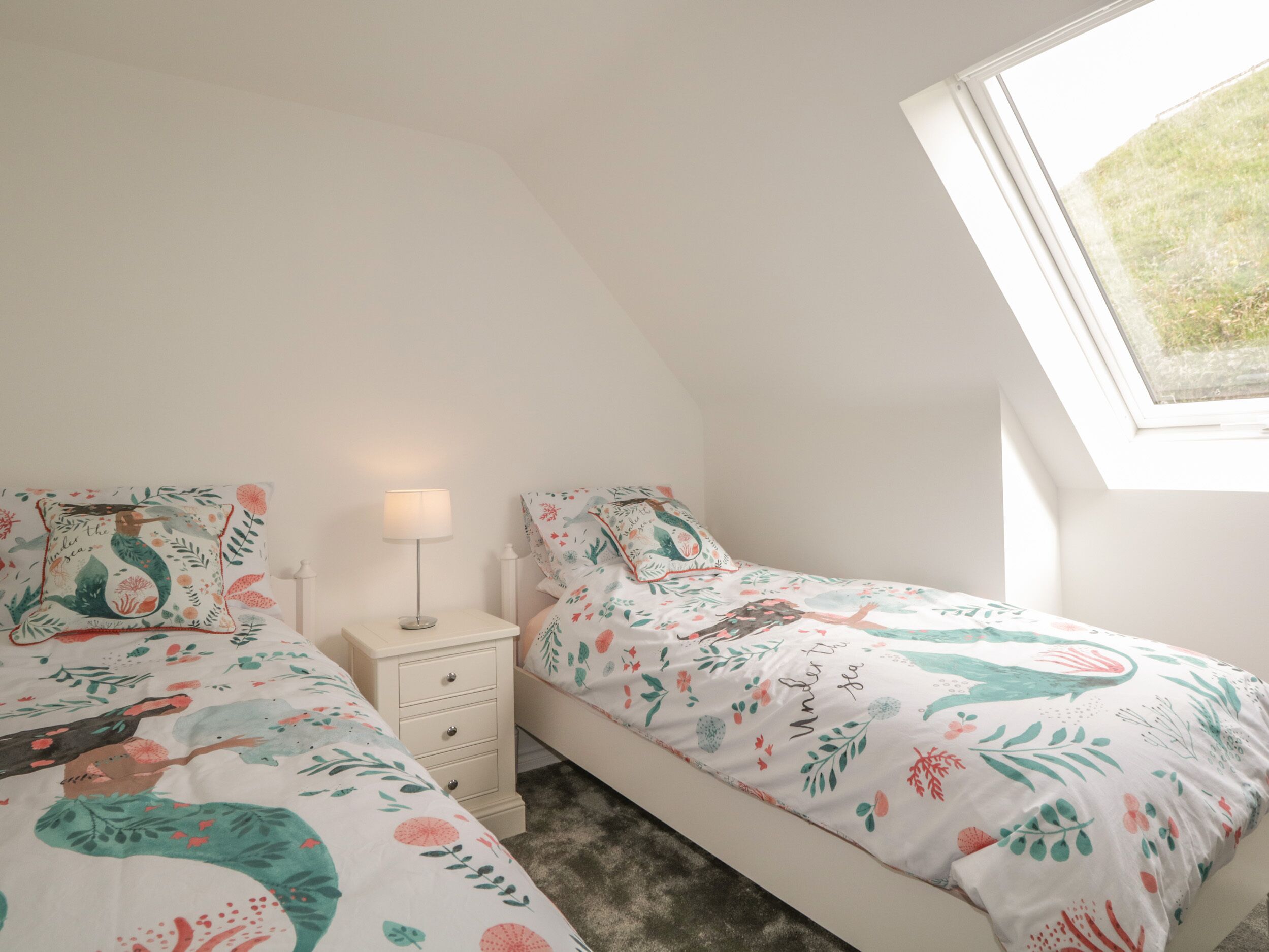 A bedroom with two beds, each with floral bedding, a bedside table with a lamp, and a window with a view of grass.