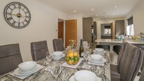 Un comedor con una mesa de mármol dispuesta para seis personas, rodeada de sillas, un gran reloj de pared y una cocina al fondo.