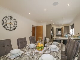 A dining area with a marble table set for six, surrounded by chairs, a large wall clock, and a kitchen in the background.