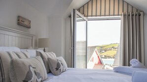 A bedroom with a bed, pillows, a lamp, and a view of a red building through an open window. - Yardarm (Salcombe)
