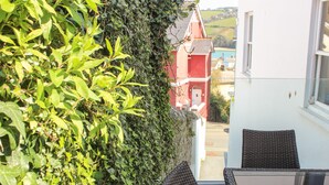 A balcony with wicker chairs and a table, overlooking a red building and a view of the countryside. - Yardarm (Salcombe)
