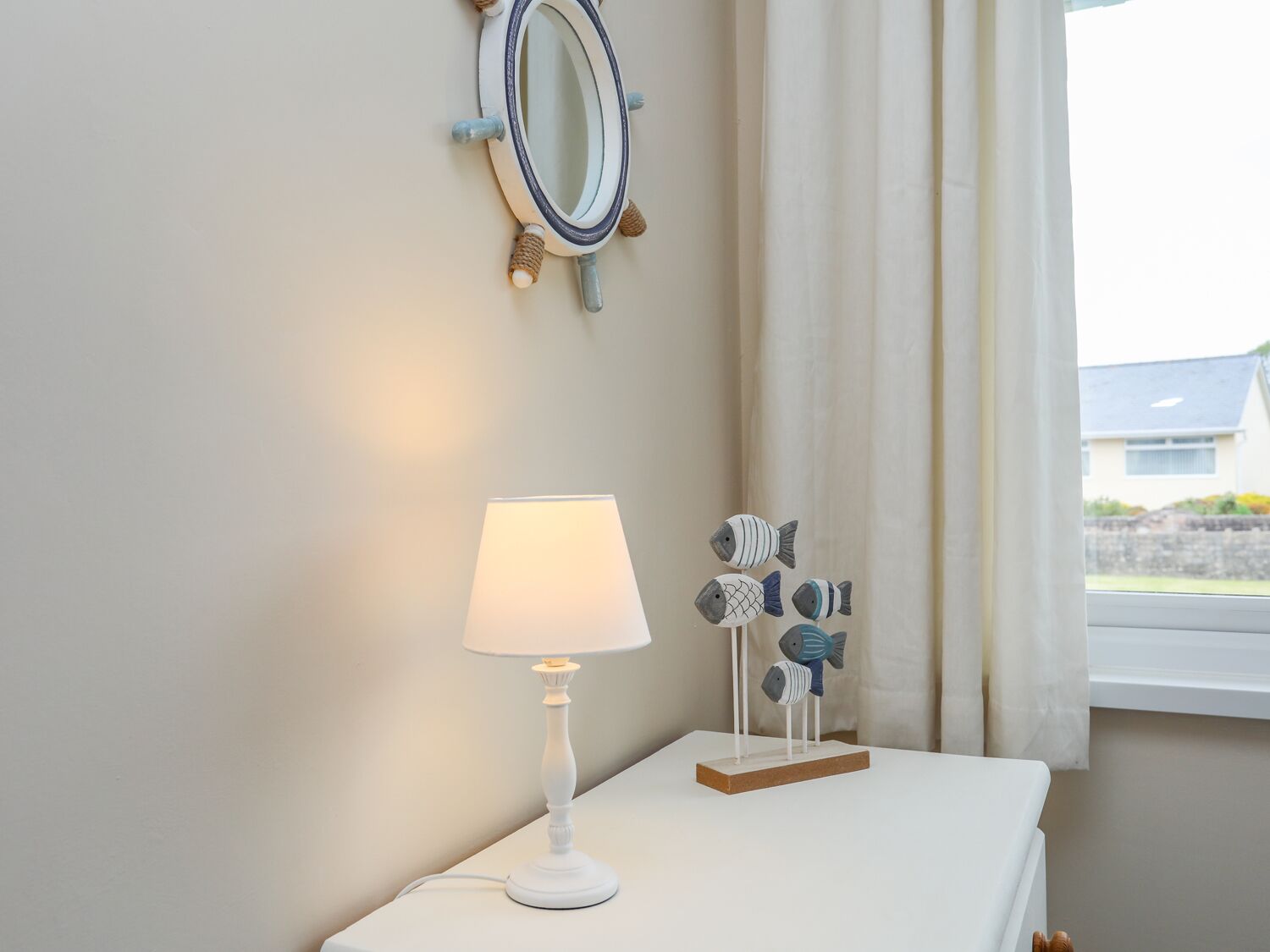 A room with a white table, a white lamp, a wall-mounted mirror, and a window with curtains.