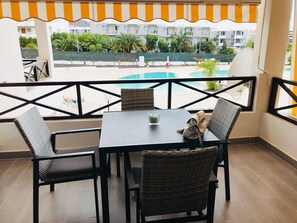 A balcony with a table and chairs, overlooking a swimming pool and greenery.