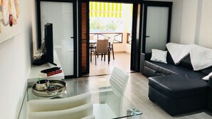 A modern living room with a glass coffee table, a black sofa, and a dining area with a table and chairs. - San Marino 2 (Los Cristianos)