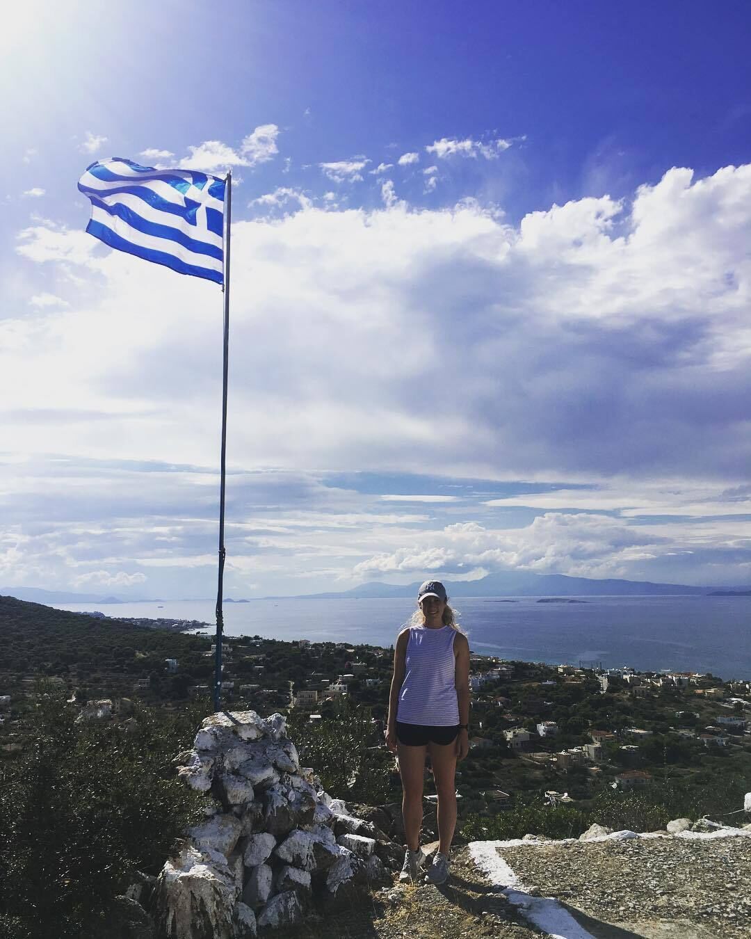 A person standing on a rocky outcrop with a flagpole bearing a flag, overlooking a coastal landscape with buildings and a view of the sea.