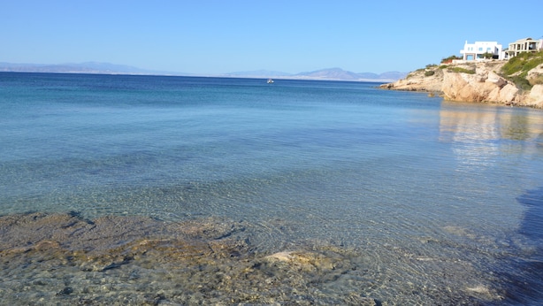 A coastal scene with clear water, rocky shore, and a white building on a cliff. - Vagia Beach Apartments (Aegina)