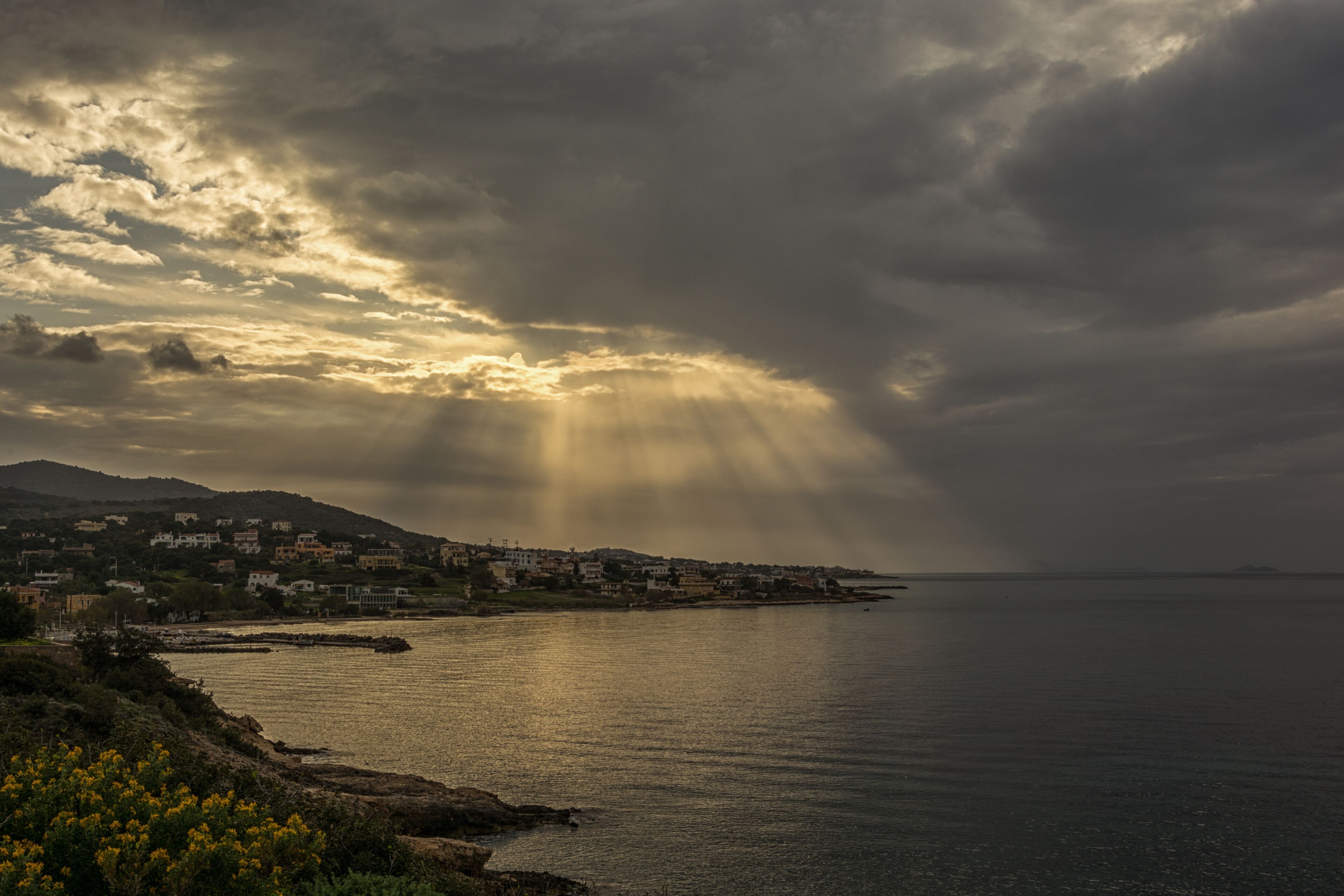 A coastal town with buildings along the shoreline, a calm sea, and a dramatic sky with sunlight breaking through clouds.