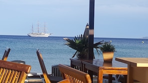 Relax in a serene outdoor seating area overlooking the sea, surrounded by lush greenery and a majestic sailing ship in the distance.