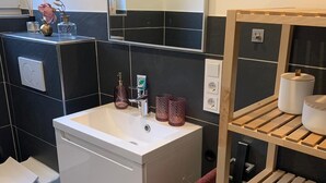 Kylpyhuoneessa on valkoinen pesuallas, mustat laatat, peili ja puinen hylly, jossa on pyyhkeitä ja kori.