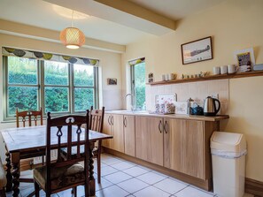A kitchen with wooden cabinets, a dining table, chairs, a hanging light fixture, a window with a view of greenery, and a shelf with kitchenware. - Elm View (Chipping Campden)