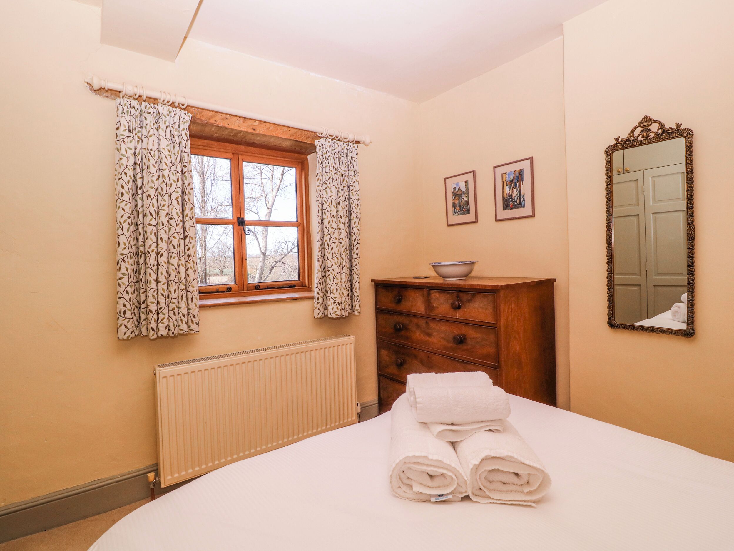 A bedroom with a bed, a dresser, a window with curtains, a radiator, and a mirror on the wall.