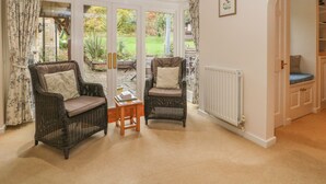 A cozy room with wicker chairs, a small table, and a view of a garden through large glass doors. - Orchard Cottage (Broadway)