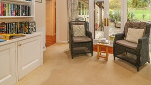 A cozy living room with wicker chairs, a bookshelf filled with books, and a view of a garden through a glass door. - Orchard Cottage (Broadway)