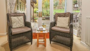 A room with two wicker chairs, a small table, and a view of a garden through glass doors. - Orchard Cottage (Broadway)