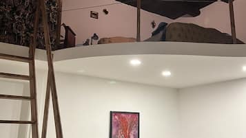 A loft bedroom with a wooden ladder, a green chair, a beige sofa, and a framed abstract painting on the wall.