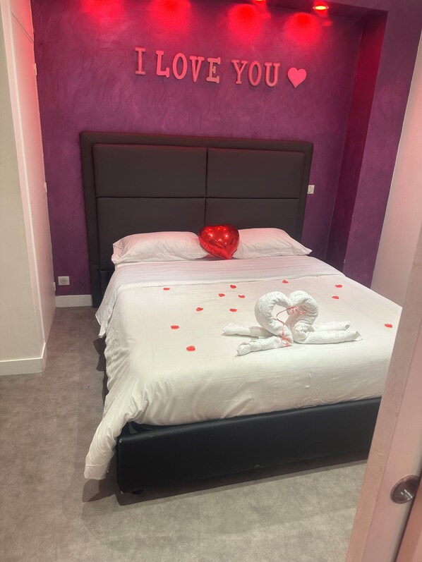 A bedroom with a bed, a wall with "I LOVE YOU" and a heart, a red heart-shaped pillow, a white swan towel, and rose petals on the bed.