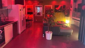 Eine moderne Küche mit Kühlschrank, Spüle und Barhocker. Ein Essbereich mit einer Couch und einer großen Topfpflanze ist ebenfalls vorhanden. Der Raum ist rot und gelb gestaltet, an der Wand befindet sich ein Schild mit der Aufschrift 'LOVE LOVE 23'.