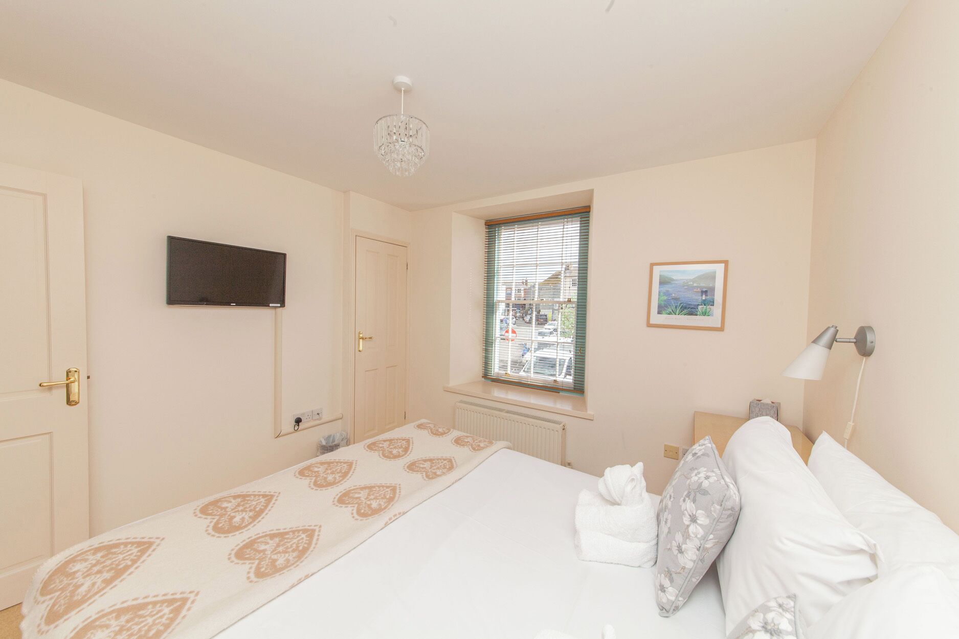 Un dormitorio con cama, televisor, ventana con persianas, radiador y un cuadro en la pared.