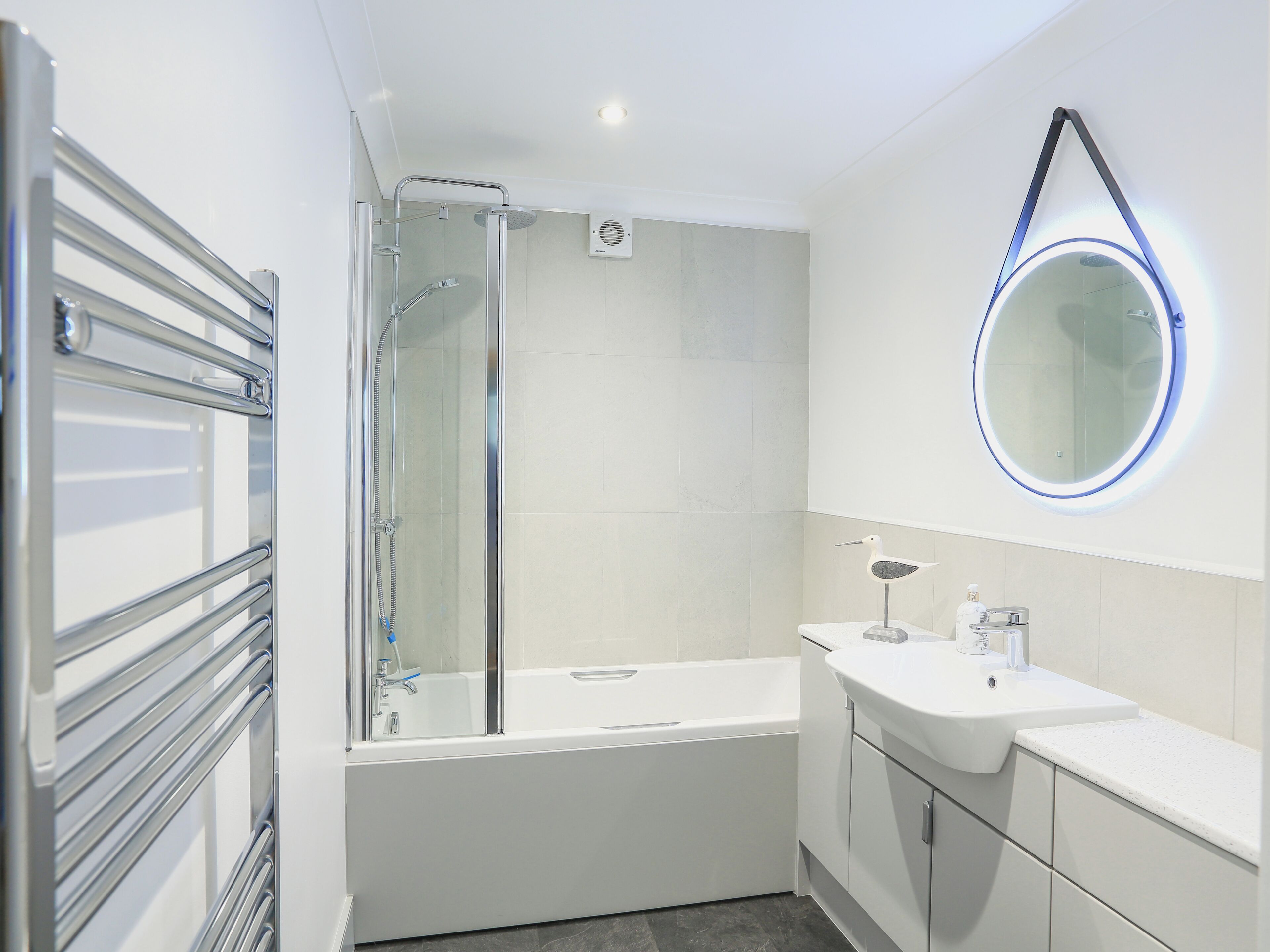 Ein modernes Badezimmer mit einer gläsernen Duschkabine, einem runden Spiegel mit blauer Beleuchtung, einem weißen Waschbecken und einer Badewanne.