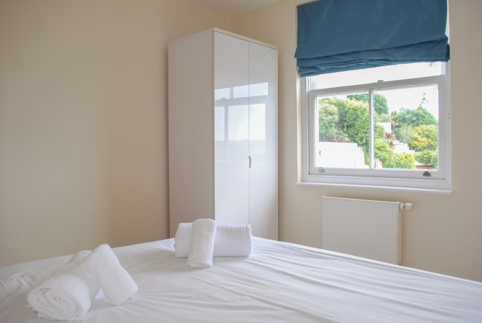 A hotel room with a bed, white towels, a wardrobe, a window with a view of greenery, and a radiator.