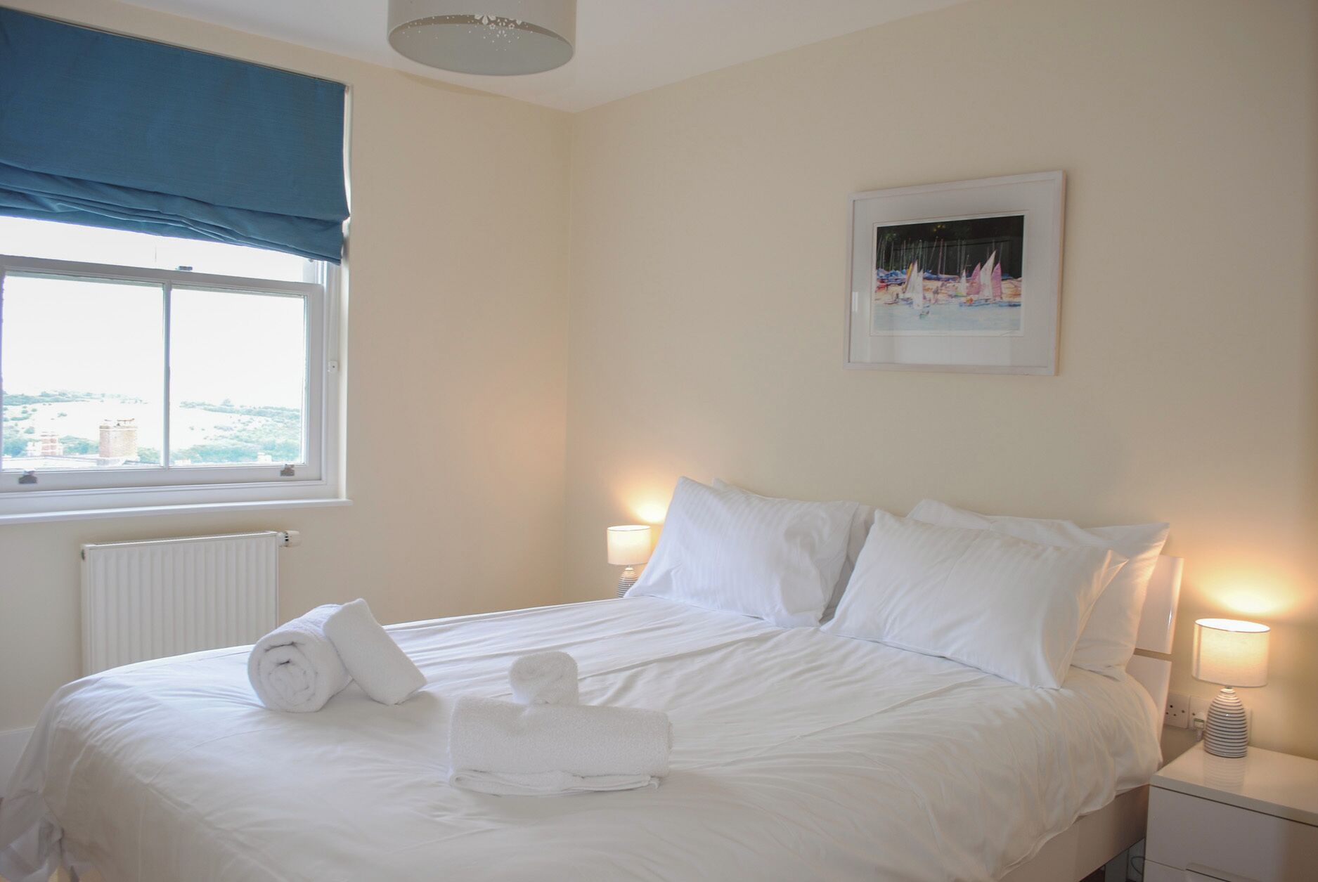 A hotel room with a bed, two bedside lamps, a window with a view, and a painting on the wall.