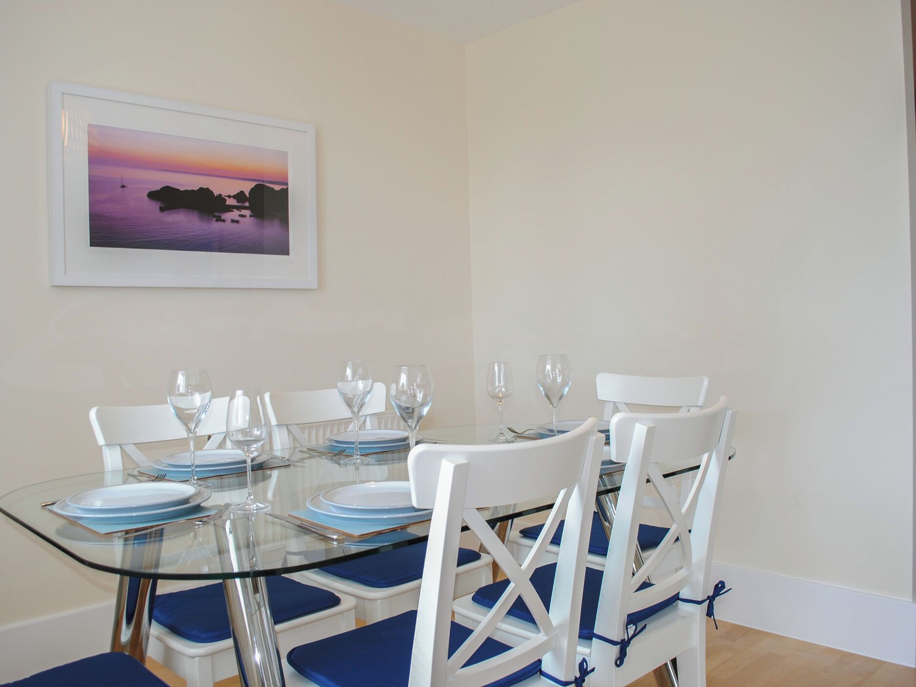 A dining area with a glass table, white chairs with blue cushions, and a framed picture of a sunset over water on the wall.