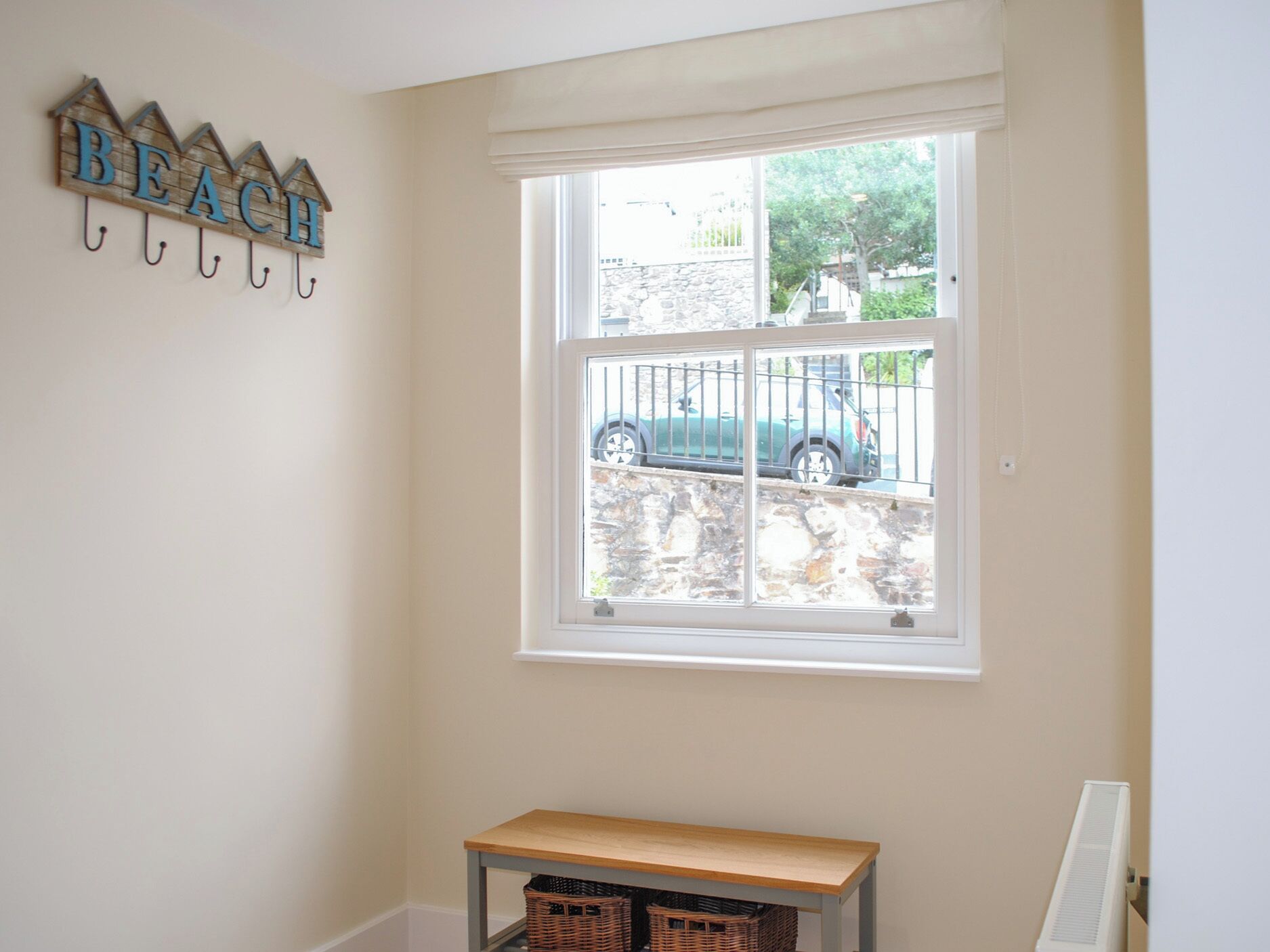 A room with a window overlooking a stone wall and parked cars. There is a wooden bench and a wall-mounted sign with the word "BEACH".
