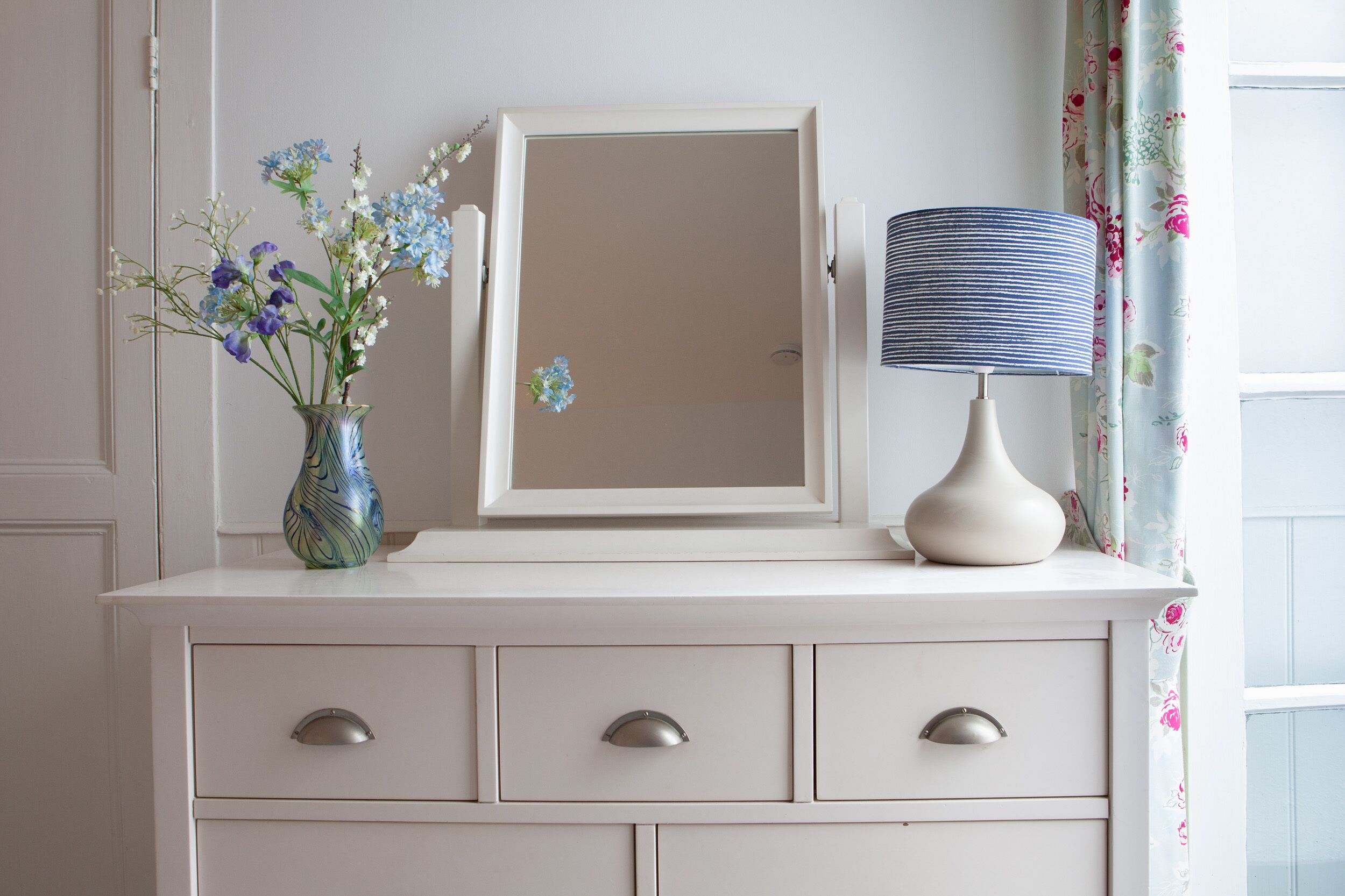 A white dresser with a mirror, a vase with flowers, and a lamp.