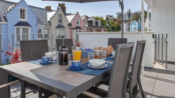 Et spisebord dækket med morgenmad, en kaffepotte og et glas appelsinjuice på en terrasse med udsigt til farverige huse.