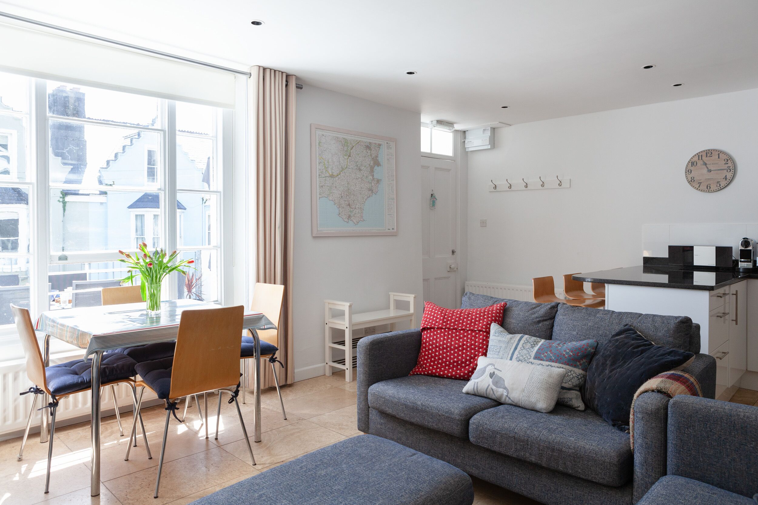 A modern living room with a grey sofa, a dining table with chairs, a large window with curtains, a map on the wall, and a kitchen area with a clock.