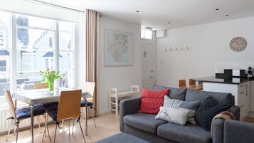 A modern living room with a grey sofa, a dining table with chairs, a large window with curtains, a map on the wall, and a kitchen area with a clock.