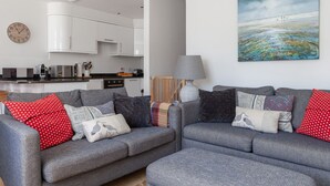 En moderne stue med en grå sofa, et maleri af en strand og et køkken i baggrunden.