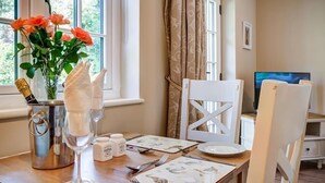Un coin repas avec une table dressée pour deux personnes, un vase de fleurs, une bouteille de champagne, des salières et des poivrières, et un téléviseur en arrière-plan.
