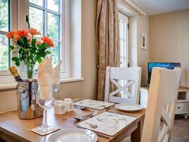 A dining area with a table set for two, a vase of flowers, a champagne bottle, salt and pepper shakers, and a television in the background.
