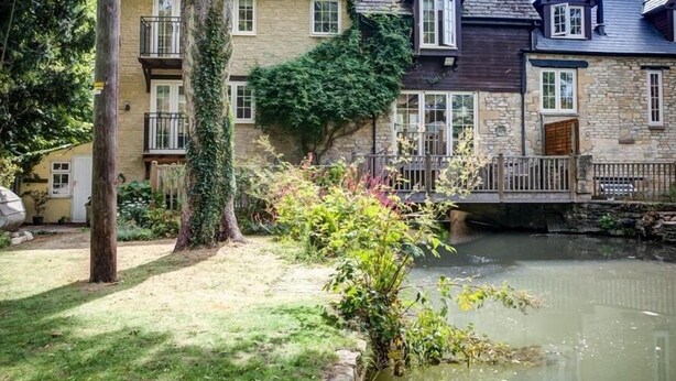 Une maison traditionnelle en pierre, avec un étang au premier plan, entourée d’une végétation luxuriante et d’une clôture en bois.
