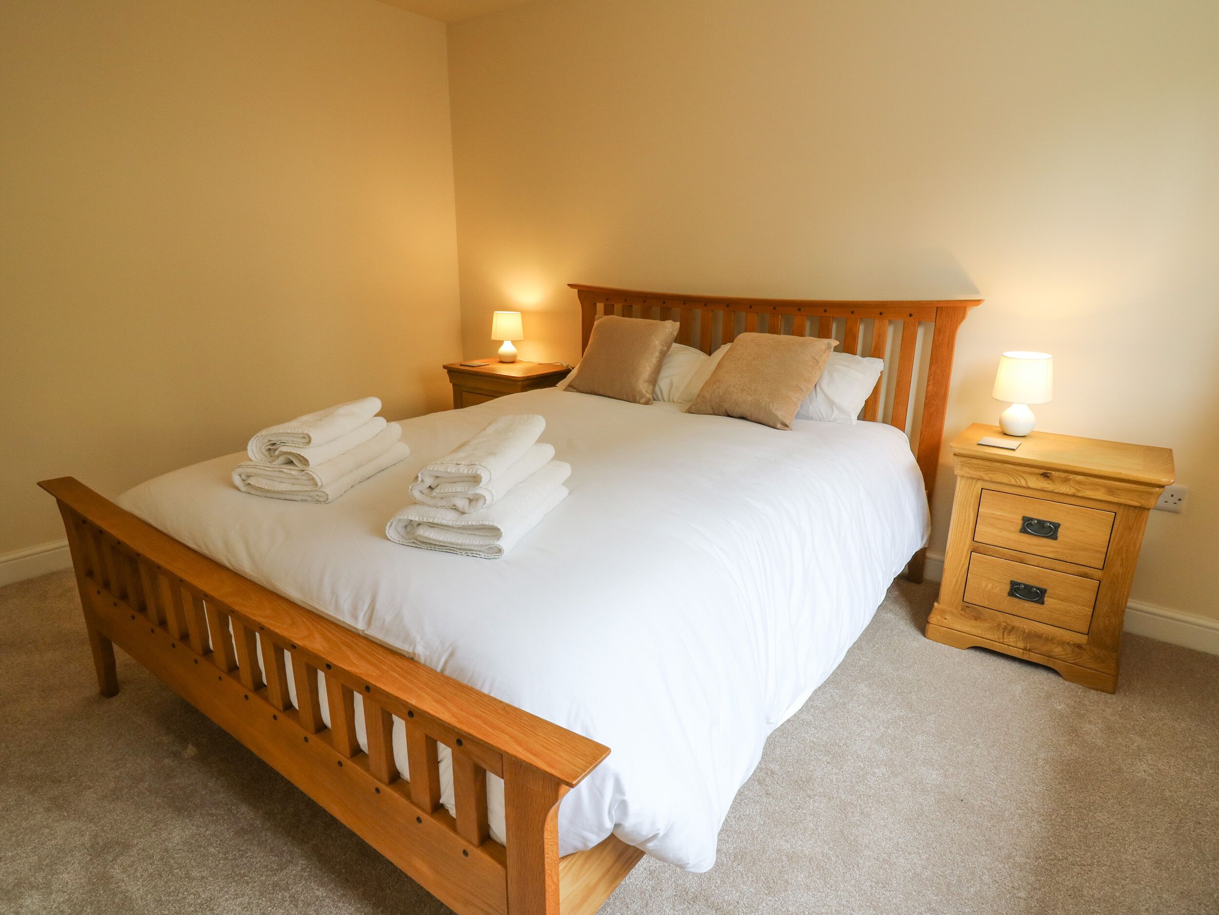 A neatly made bed with white linens and folded towels, wooden headboard, two bedside tables with lamps, and a carpeted floor.