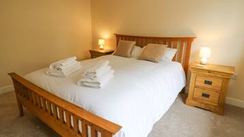 A neatly made bed with white linens and folded towels, wooden headboard, two bedside tables with lamps, and a carpeted floor.