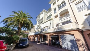 Un immeuble résidentiel à plusieurs étages, doté de balcons, d’un espace de stationnement couvert et d’un palmier.