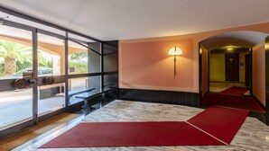 Un hall d’hôtel avec un sol en marbre, une zone recouverte d’un tapis rouge, des portes vitrées donnant sur l’extérieur et une lampe fixée au mur.