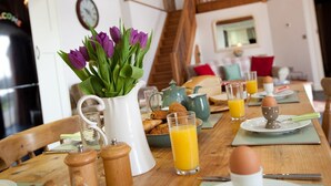 Une table de petit-déjeuner avec du jus d’orange, des œufs et des viennoiseries, un pichet blanc orné de fleurs violettes, et un escalier en bois en arrière-plan.