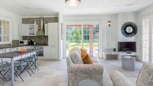 Une cuisine moderne avec des meubles blancs, un coin repas comprenant une table et des chaises, un canapé, un téléviseur et une grande fenêtre donnant sur un paysage verdoyant.