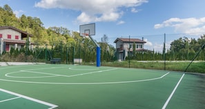 An outdoor basketball court with a chain-link fence, trees, and houses in the background.