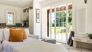 Une chambre avec un grand lit, un canapé, une vue sur un jardin par une porte-fenêtre coulissante, et une cuisine à l’arrière-plan.
