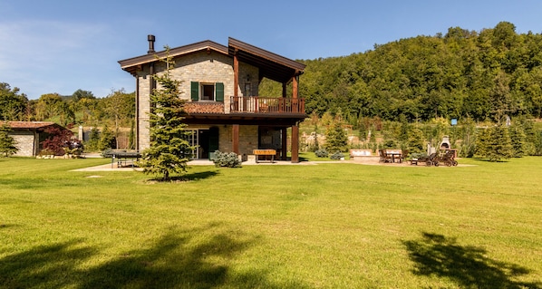 A two-story stone house with a balcony, surrounded by a well-maintained lawn and trees.