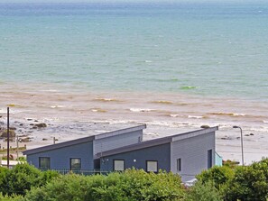 A modern building with a flat roof, located near a beach with visible waves. - Ty Glyn (Criccieth)