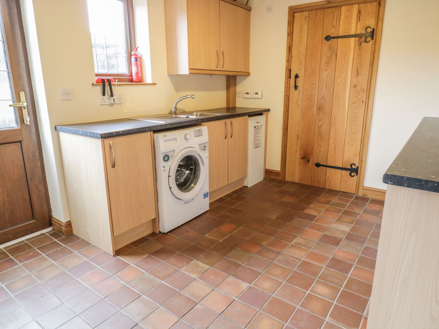A compact kitchen with a washing machine, wooden cabinets, a tiled floor, and a wooden door.