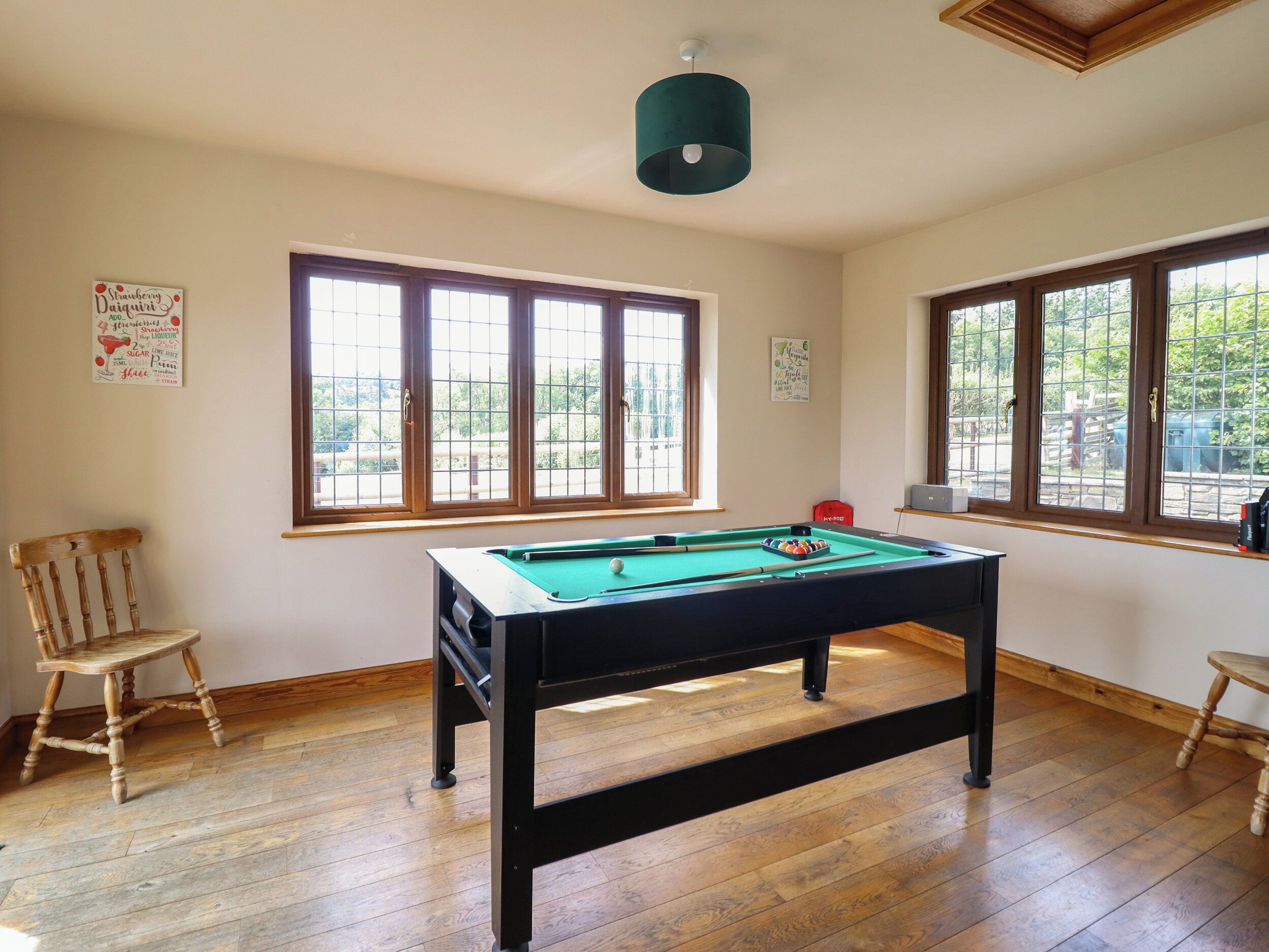 A pool table in a room with wooden flooring, large windows, and a ceiling light fixture.