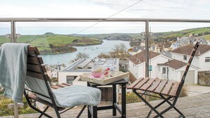 Ein Balkon mit zwei Holzstühlen, einem kleinen Tisch mit Tasse und Untertasse und Blick auf einen Fluss und Häuser in der Ferne.